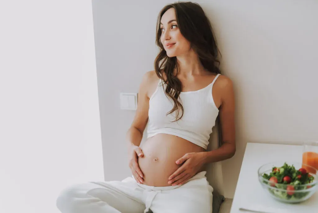 Pretty calm mama holding hands on stomach and sitting on kitchen chair inside light flat